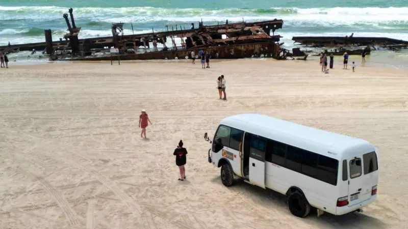 Fraser Island 2 Day Tour van, tourists on sandy beach beside iconic rusted shipwreck, scenic ocean view, top travel experience.