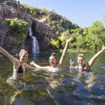 Three happy women in swimming costumes on a Kakadu Tour camping trip raise their arms by a stunning waterfall and crystal-clear natural pool.