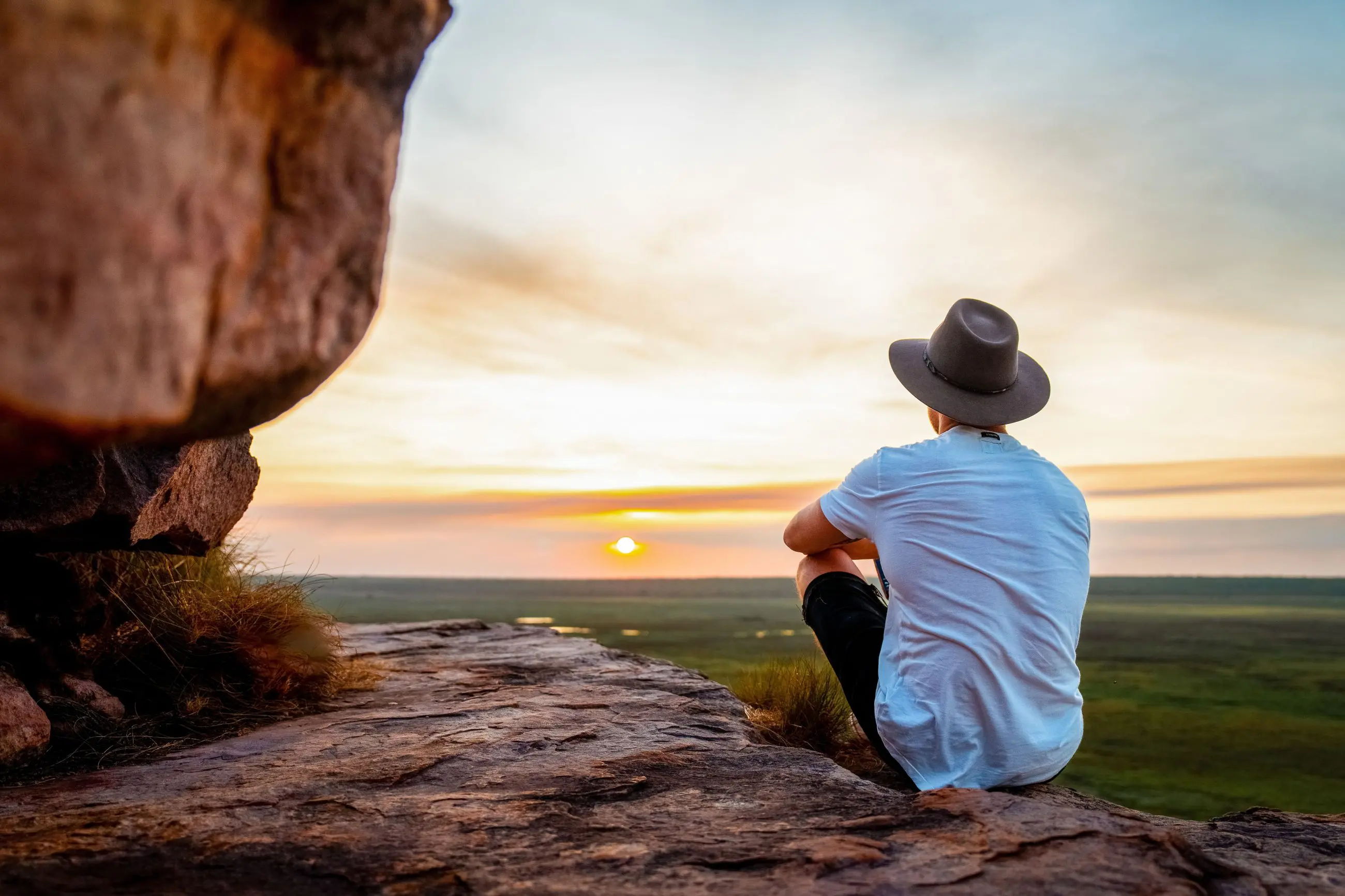 Traveller wearing a hat sits on a rugged rocky ledge, admiring Kakadu's breathtaking sunset over vast, dramatic cloudy landscapes.