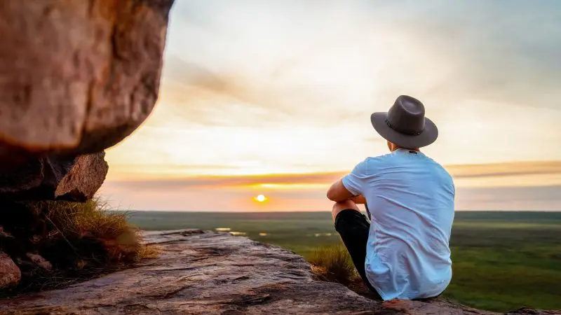 Traveller wearing a hat sits on a rugged rocky ledge, admiring Kakadu's breathtaking sunset over vast, dramatic cloudy landscapes.