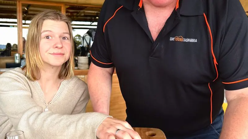 Cheerful man presenting gourmet cheese cubes on a wooden platter during Huon Valley Safaris Private Day Tour at restaurant table.