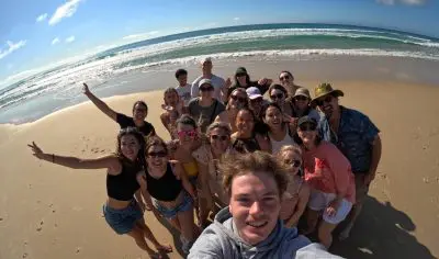 Friends pose for a bright selfie on an East Coast Explorer tour, smiling with rolling ocean waves and sunny skies in the background.