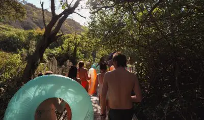 Group of holidaymakers in swimming costumes stroll along a sunlit promenade, carrying lilos on their East Coast Explorer beach holiday.