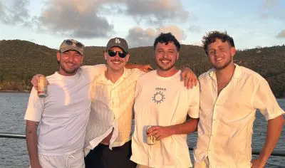 Four men in white outfits enjoy drinks and smile on a boat tour, East Coast Explorer, with lush green hills and sparkling water behind them.
