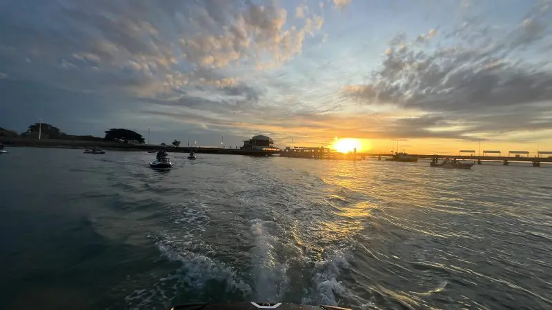 A jet ski speeds across the water towards a pier at Honey Rider Sunrise, beneath dramatic clouds and a glowing sun on the horizon.