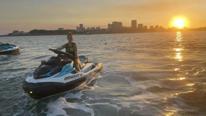 Adventurer jet skiing at sunset on the Golden Eye Sunset Tour, vibrant city skyline in background, glowing water reflecting orange sky.