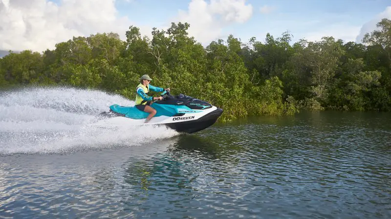 Person in life jacket speeding on jet ski James Bond style over a serene lake, surrounded by lush green trees and vibrant bushes.