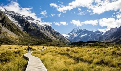 Adventurers hike a scenic wooden boardwalk through lush New Zealand fields towards majestic snow-capped mountains under clear skies.