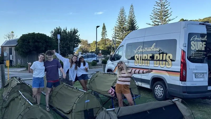 Four travellers by tents and the iconic Australian Surf Bus on a 6-Day Sydney to Brisbane Surf Trip beneath clear blue skies.