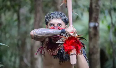 A person in vibrant traditional dress with a painted face presents a cultural gesture at Waitangi Treaty Grounds forest, New Zealand.