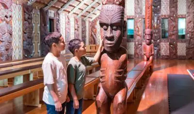 Two boys inside the historic carved wooden meeting house at Waitangi Treaty Grounds, interacting with a large traditional Māori statue.