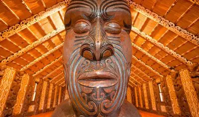 Detailed close-up of intricately carved wooden Māori statue at Waitangi Treaty Grounds, inside meeting house with ornate ceiling décor.
