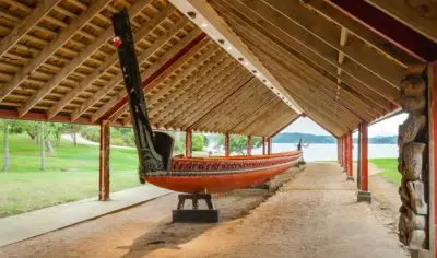 A historic Viking ship rests under a wooden shelter at Waitangi Treaty Grounds, set beside lush grass and scenic waterfront views.