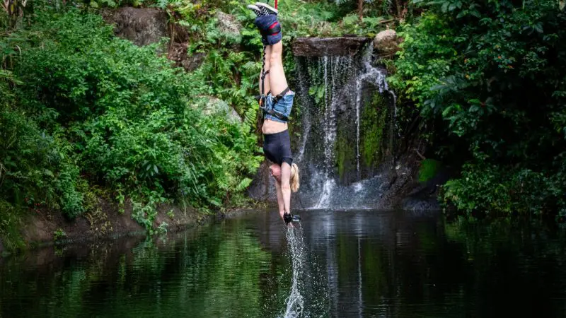 Thrill-seeker bungee jumping upside down at Roof Jump Skypark, splashing water with hands amid lush green forest and scenic waterfall.