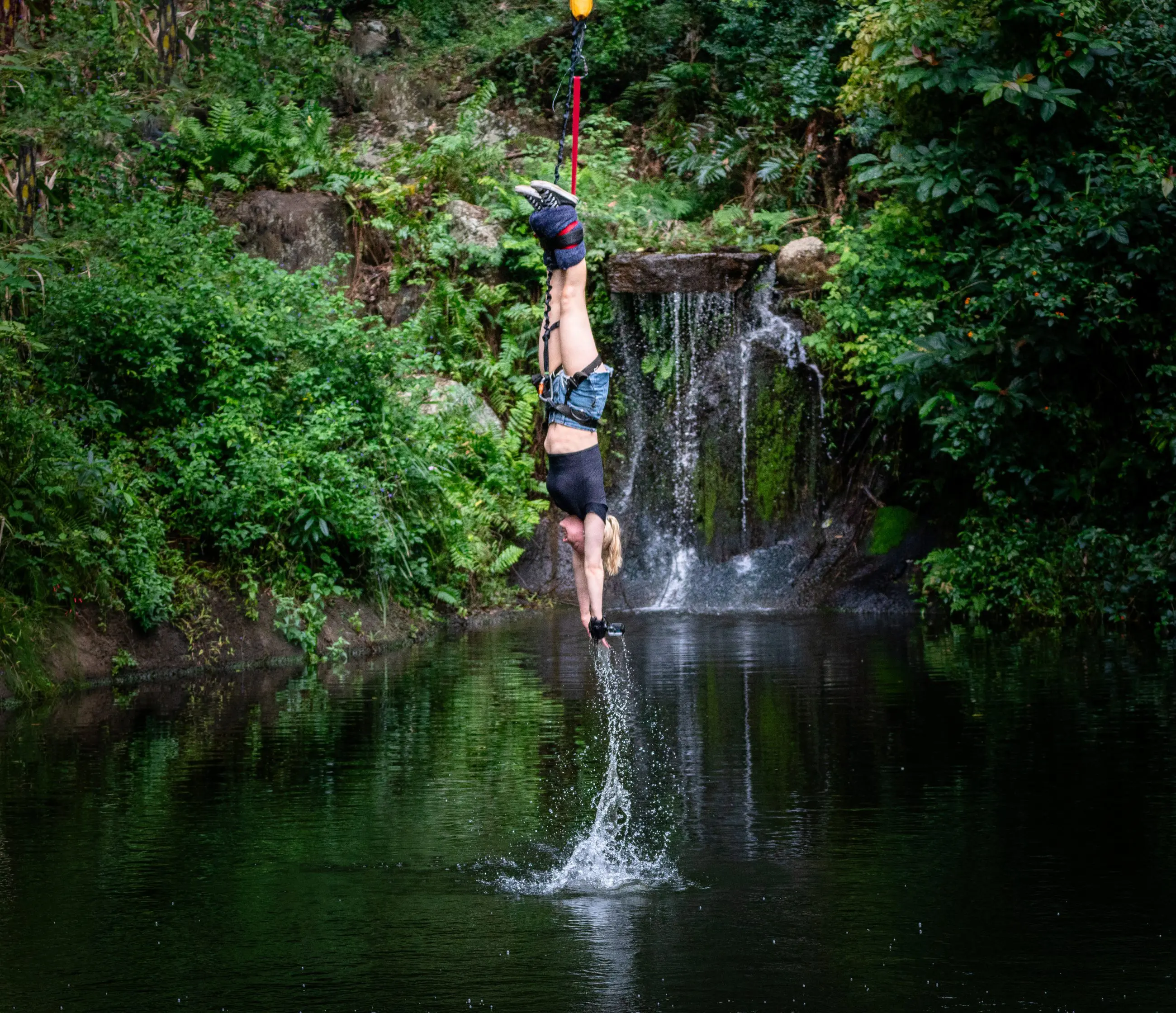 Experience the thrill of a Bungee Jump Swing Combo as a person dangles upside down above a scenic pond near a cascading waterfall.