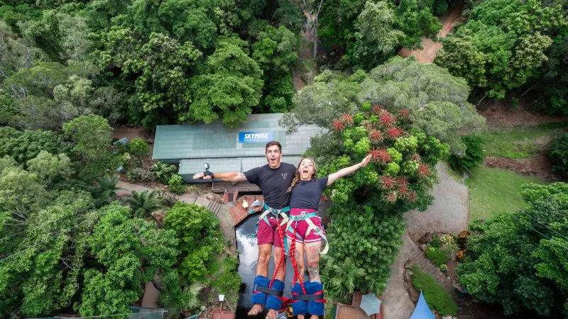 Two thrill-seekers in safety harnesses get ready to bungee jump at Australia’s Only Bungy Skypark, soaring above a lush green rainforest.