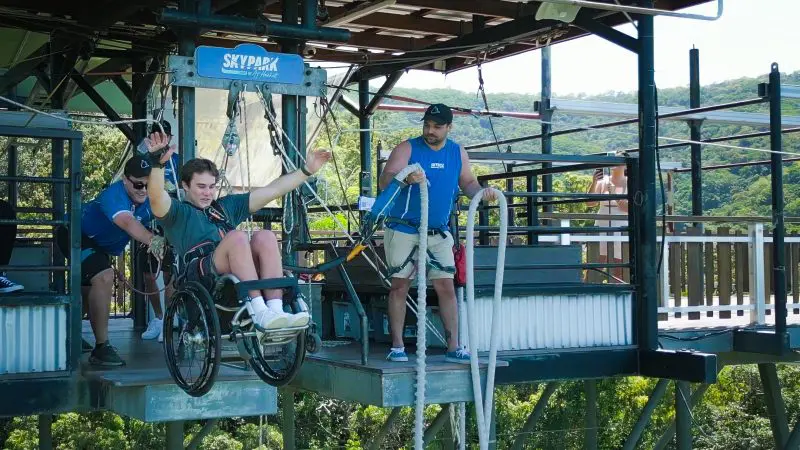 At Australia’s Only Bungy Skypark, a wheelchair user gets assistance to bungy jump above lush green forest treetops.