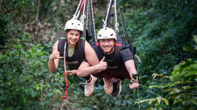 Two adventurers zipwire side by side above a vibrant green forest, smiling and giving thumbs up—ideal for a Bungee Jump Swing Combo.