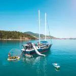 Experience The Great Escape Australia: 28-Day Semi-Guided yacht with water slides, anchored in crystal-clear waters amid small boats.