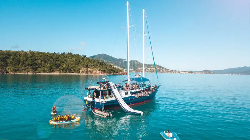 Experience The Great Escape Australia: 28-Day Semi-Guided yacht with water slides, anchored in crystal-clear waters amid small boats.