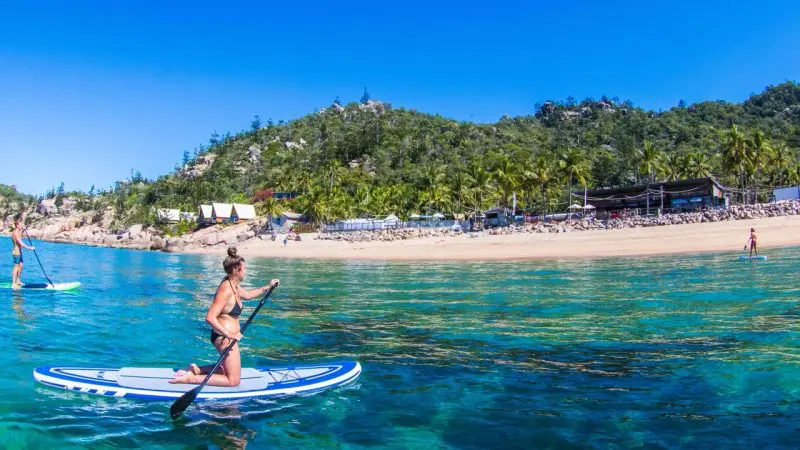 Woman kayaking on crystal-clear blue water beside lush palm trees, evoking The Great Escape 21-Day Trip from Brisbane to Cairns.