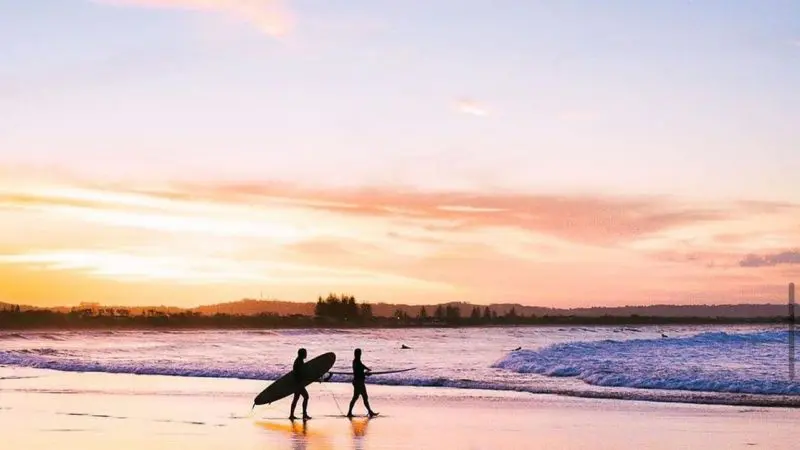 Two surfers carrying boards stroll along the beach at sunset, inspired by The Great Escape Australia 28 Day Semi-Guided adventure tour.