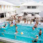 People swim and play with beach balls at a lively, sunny Australia group trip resort with white buildings.