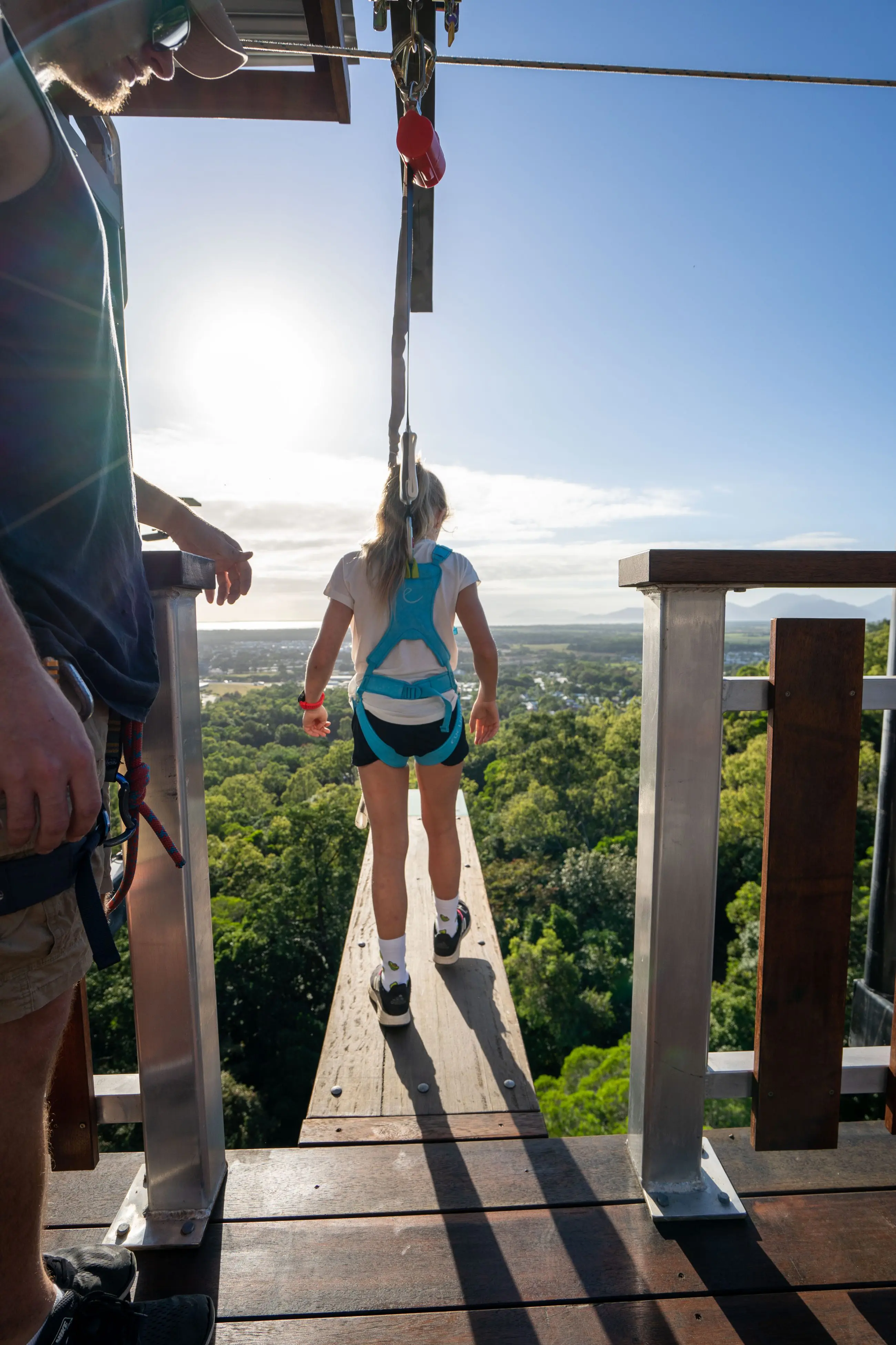Adventurer in safety harness readies for thrilling zip wire ride at Walk The Plank Skypark, above vibrant green landscape on sunny day.
