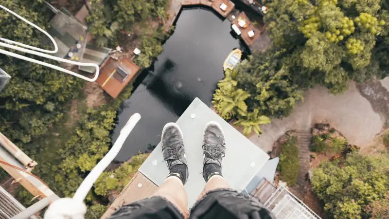 Close-up of feet at the edge of Walk The Plank Skypark’s high dive, overlooking a sparkling pool, lush trees, and city buildings below.