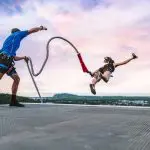 Thrill-seeker leaps for a Skypark Roof Jump at sunset as safety instructor fastens rope, highlighting extreme adventure sports experience.
