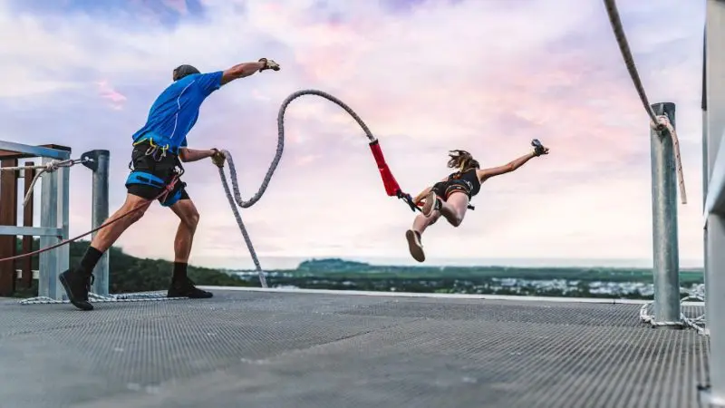Thrill-seeker leaps for a Skypark Roof Jump at sunset as safety instructor fastens rope, highlighting extreme adventure sports experience.