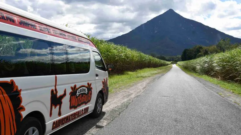 Creek To Coral Mission Beach Day Tour van parked beside lush tall grass on a scenic rural road with mountains and cloudy sky backdrop.