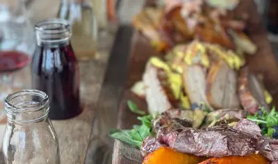 Gourmet wooden platter with grilled meats, roasted squash, artisan sandwiches on rustic table; empty glass jars accent the scene.