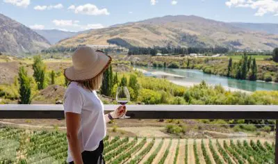 A woman in a sunhat savours premium wine on a luxury boutique wine tour, overlooking scenic vineyards, majestic mountains, and winding river.