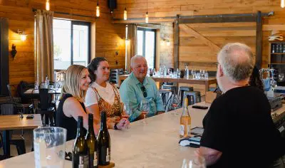 Three guests savour a Classic Wine Tour, smiling and interacting with the barman at a charming rustic winery tasting bar counter.