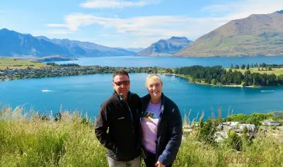 Happy couple on a lush green hill in Queenstown with panoramic lake and mountain views under a clear blue sky, perfect travel scene.