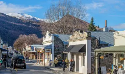 Visitors walk along a vibrant Queenstown street lined with cafes and boutique shops, framed by stunning mountains—ideal for tours.