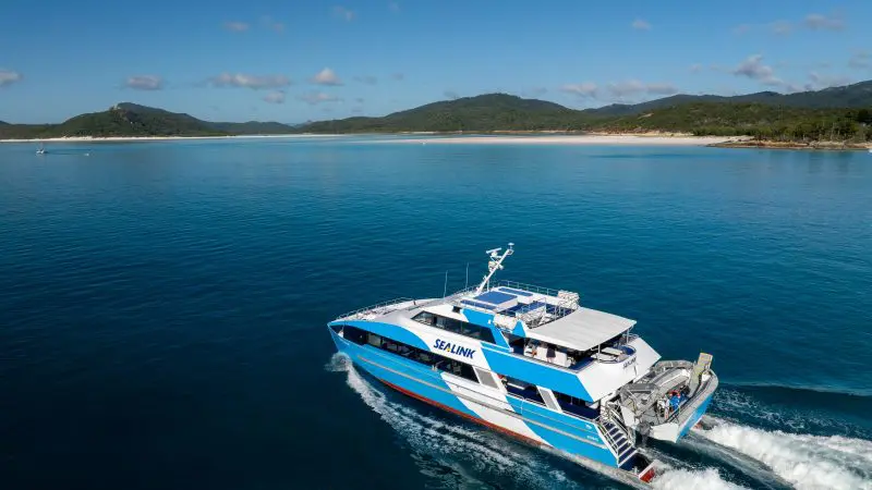 SeaLink ferry on the Whitehaven Day Tour sails smoothly over crystal blue waters, backed by lush green hills and scenic coastline views.