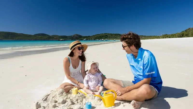 Family with toddler playing with sand toys on Purely Whitehaven Day Tour, beside pristine blue water and lush green hills, Australia.