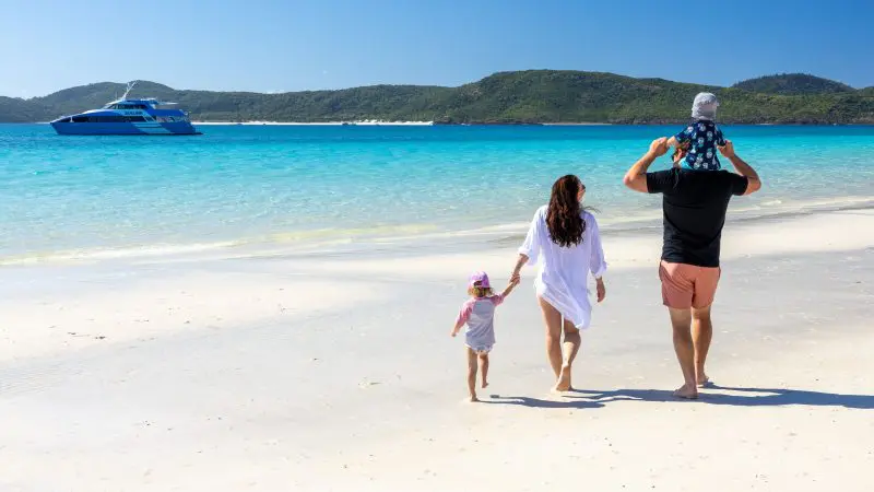 A smiling family of four walks along Whitehaven Beach under clear skies, with vibrant turquoise waters and lush green hills behind them.