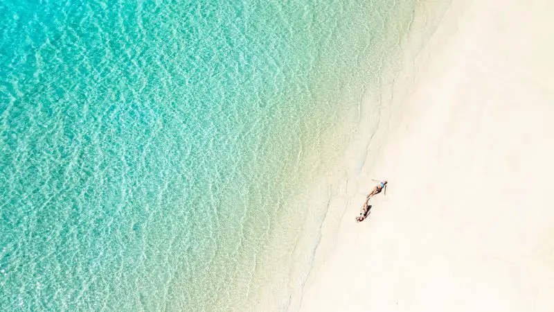 Stunning aerial view of two tourists on a Purely Whitehaven Day Tour relaxing on pristine white sand beside crystal-clear blue water.
