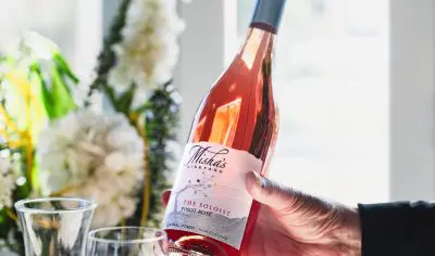 A hand displays Misha’s Vineyard The Soloist Pinot Rosé wine bottle by a glass and fresh flowers, illuminated in vibrant sunlight.