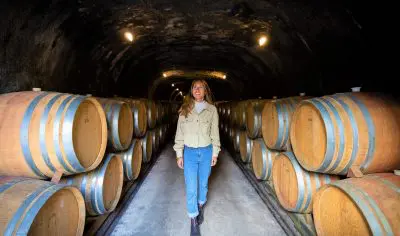 Elegant woman strolling through vintage wine cellar tunnel lined with oak barrels during guided wine tour, low ambient lighting.