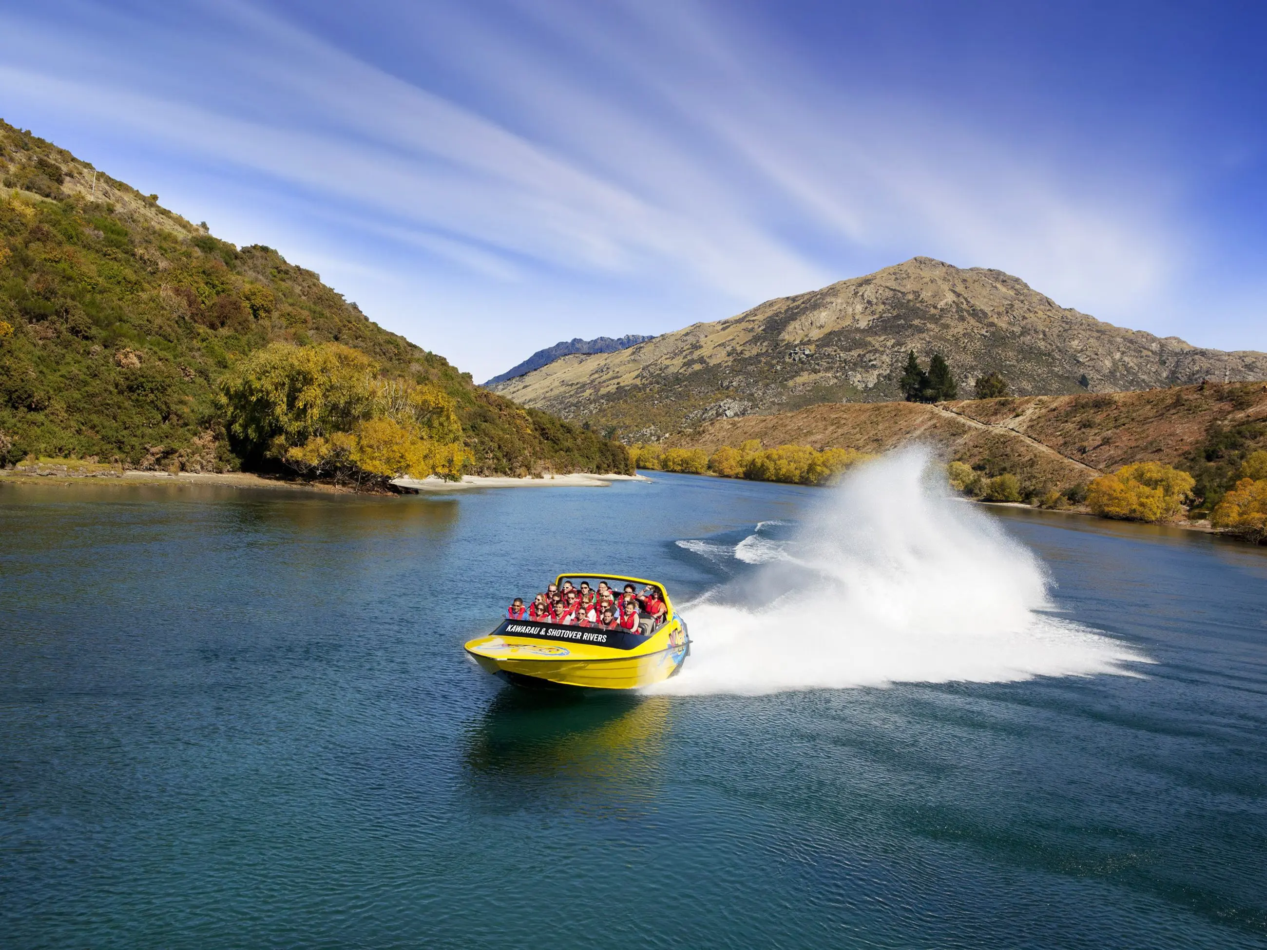 Experience the thrill as a yellow jet boat filled with excited passengers speeds across a vibrant blue river, spray erupting, lush hills and green trees behind.