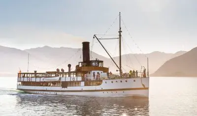 Experience a scenic wine tour aboard a historic steamship on a tranquil lake, with panoramic mountain views beneath clear sunny skies.