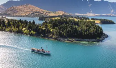 A scenic cruise boat glides on a turquoise lake beside a lush forested island, framed by majestic mountains and blue skies.