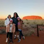 Family of four at Uluru Red Centre at sunset, standing on iconic red earth with golden grasses and clear blue skies in the Australian Outback.