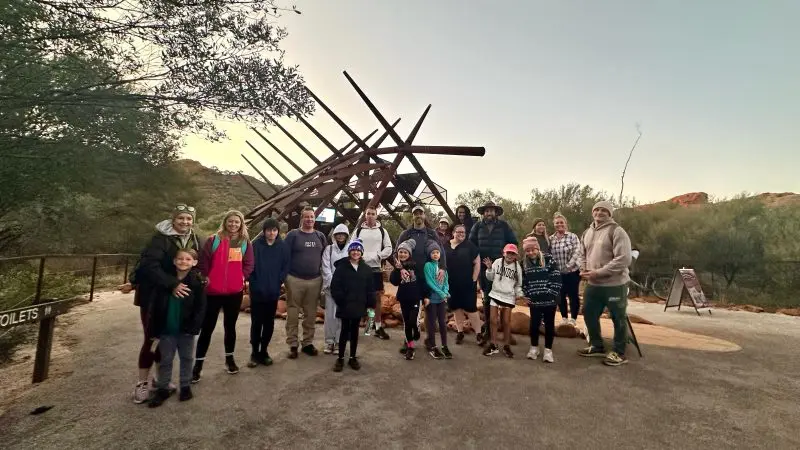 Family group enjoys the Uluru Red Centre Adventure at dusk, posing by native trees and a wooden sculpture in the scenic outback.