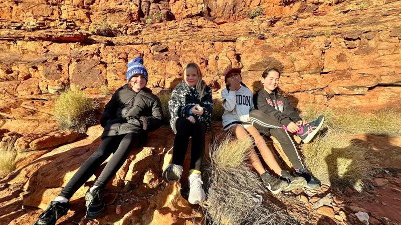 Four smiling children enjoy a family adventure sitting on Uluru’s iconic red rocks, dressed for the sunny Australian Outback experience.
