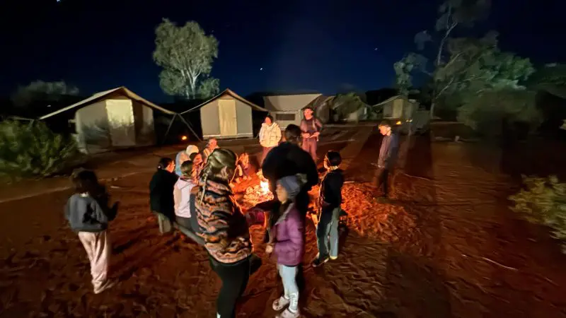 Travellers gather around a glowing campfire at night beside tents in Uluru’s Red Centre, experiencing an epic Yulara adventure.